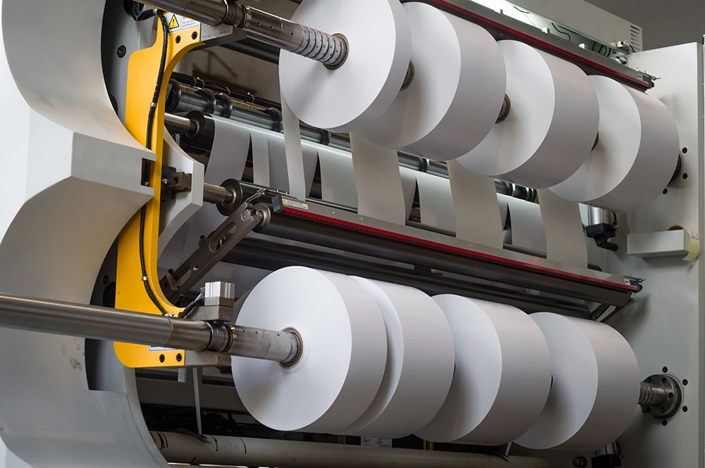 Large industrial machine unwinding and cutting wide rolls of white paper into narrower rolls in an Adhesives Research manufacturing setting.
